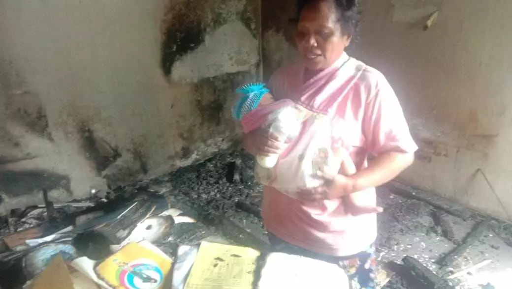 Nona elwuar sambil. Menggendong anak melihat kondisi rumah orang tua yang terbakar dan rusak